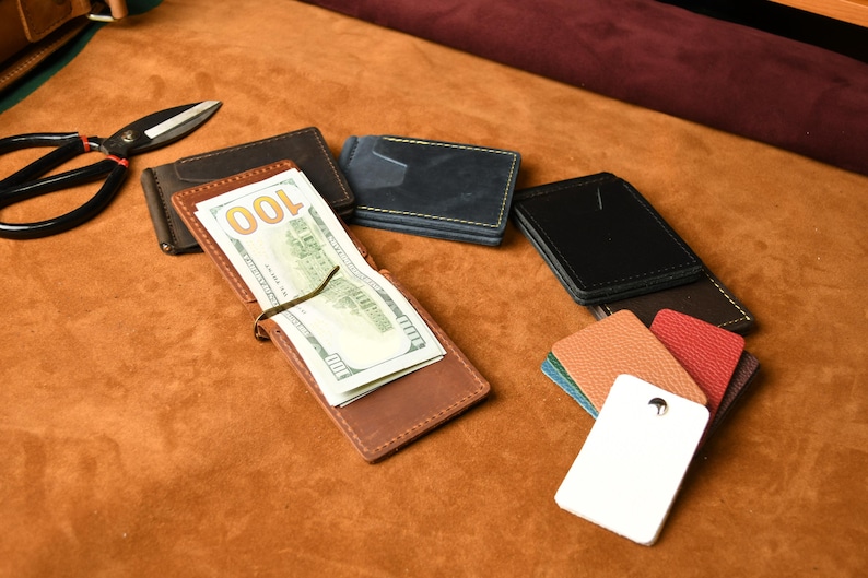 Puede incluir: Una colecci&oacute;n de carteras con clip para billetes de cuero en varios colores, incluyendo marr&oacute;n, negro y azul, con una cartera marr&oacute;n que contiene un billete de cien d&oacute;lares. Se incluyen unas tijeras de metal negro y muestras de cuero texturizado.