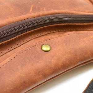 May include: Close-up of a brown leather bag with a zipper and a brass-colored snap. The leather has a distressed look with visible stitching and a textured surface. The zipper is dark brown.