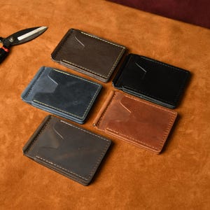 Puede incluir: Cinco carteras rectangulares de cuero con clip para billetes en marr&oacute;n oscuro, negro, azul marino, tostado y gris oscuro, cada una con un clip de metal y ranura para tarjetas. Se ven unas tijeras de metal negro y muestras de cuero de colores.
