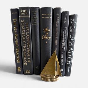 May include: A row of seven black books with gold lettering on the spines, including titles like The Sea Rover's Practice and Seawitch, arranged behind a small, triangular brass sailboat figurine with a textured base.