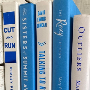 May include: A bookshelf with a row of five books. The books are white with blue titles. The titles are "Outliers", "The Roxy Letters", "Talking to Robots", "The Sisters of Summit Avenue", and "Cut and Run".