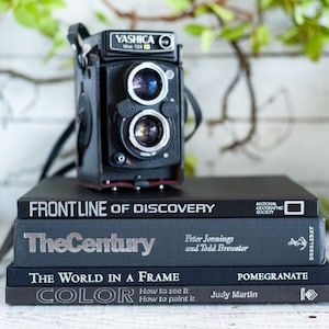 May include: A stack of four black hardcover books with silver lettering. The top book is titled "Frontline of Discovery" and has the National Geographic Society logo. The second book is titled "The Century" and has the author names Peter Jennings and Todd Brewster. The third book is titled "The World in a Frame" and has the author name Judy Martin. The bottom book is titled "Color" and has the text "How to see it, How to paint it". A vintage black camera sits on top of the books.
