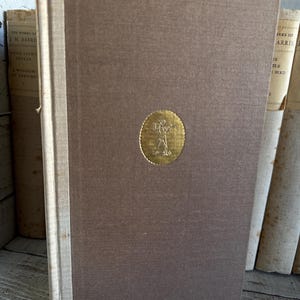 Puede incluir: Un libro de tapa dura marr&oacute;n con un emblema dorado. El lomo es de color beige claro. Otros libros son visibles en el fondo. El libro forma parte de una colecci&oacute;n de libros antiguos.