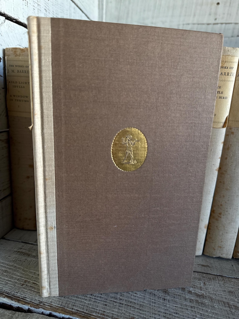 Puede incluir: Un libro de tapa dura marr&oacute;n con un emblema dorado. El lomo es de color beige claro. Otros libros son visibles en el fondo. El libro forma parte de una colecci&oacute;n de libros antiguos.