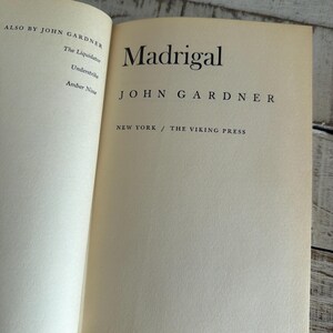 May include: An open book displaying the title "Madrigal" by John Gardner. The book has off-white pages with black text. Published by The Viking Press, New York. Other works by the author are listed on the left page.