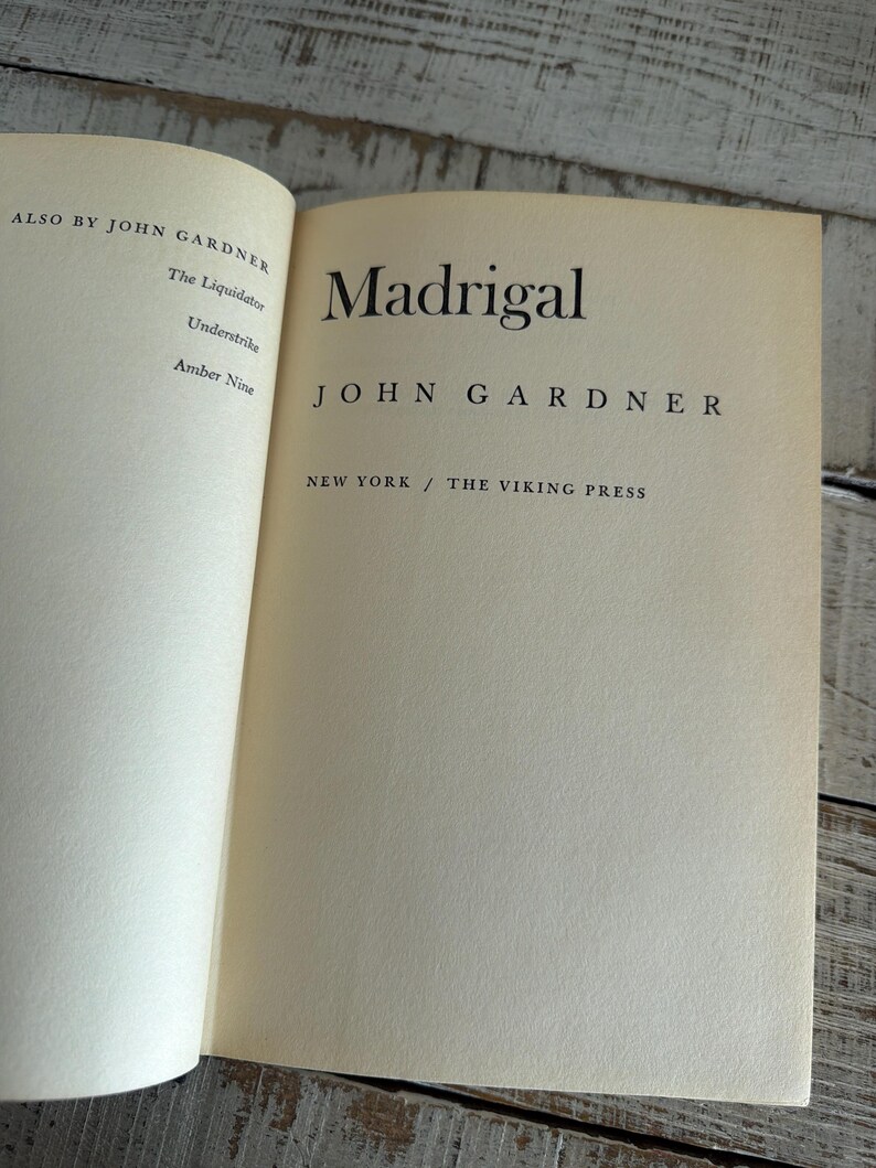 May include: An open book displaying the title "Madrigal" by John Gardner. The book has off-white pages with black text. Published by The Viking Press, New York. Other works by the author are listed on the left page.