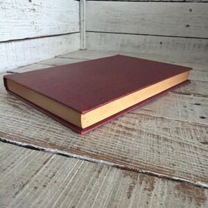 Puede incluir: Un libro de tapa dura con una cubierta burdeos y bordes de p&aacute;gina dorados. El libro est&aacute; tumbado sobre una superficie de madera envejecida. La cubierta tiene una textura.