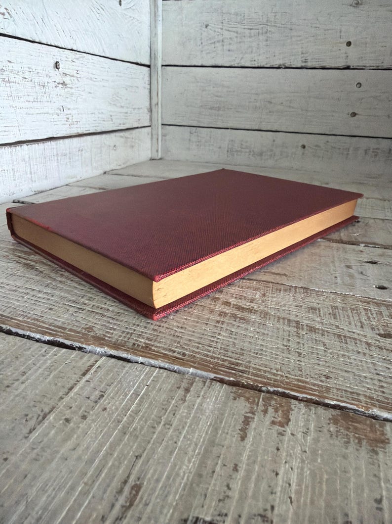 Puede incluir: Un libro de tapa dura con una cubierta burdeos y bordes de p&aacute;gina dorados. El libro est&aacute; tumbado sobre una superficie de madera envejecida. La cubierta tiene una textura.