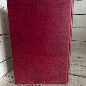 Puede incluir: Un libro de tapa dura con una cubierta de color rojo oscuro. La cubierta tiene una apariencia texturizada y muestra un ligero desgaste en el borde inferior. El lomo es visible y el libro est&aacute; colocado sobre una superficie de madera blanca.