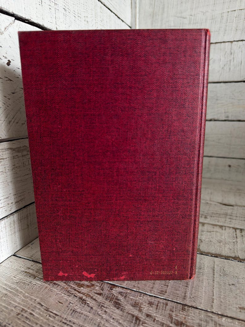 Puede incluir: Un libro de tapa dura con una cubierta de color rojo oscuro. La cubierta tiene una apariencia texturizada y muestra un ligero desgaste en el borde inferior. El lomo es visible y el libro est&aacute; colocado sobre una superficie de madera blanca.