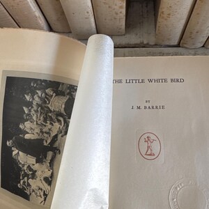 Puede incluir: Libro abierto con el t&iacute;tulo "THE LITTLE WHITE BIRD" de J.M. Barrie. La portada es de color crema con texto e ilustraciones en rojo y negro. Las p&aacute;ginas est&aacute;n envejecidas y el libro est&aacute; rodeado de otros libros.