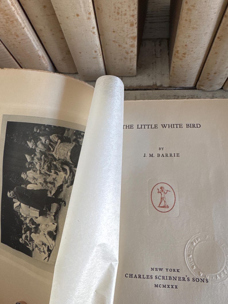 Puede incluir: Libro abierto con el t&iacute;tulo "THE LITTLE WHITE BIRD" de J.M. Barrie. La portada es de color crema con texto e ilustraciones en rojo y negro. Las p&aacute;ginas est&aacute;n envejecidas y el libro est&aacute; rodeado de otros libros.