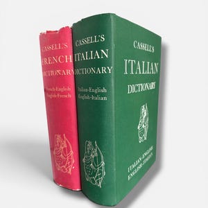 Op de afbeelding: Twee vintage woordenboeken: "Cassell's French Dictionary" in rood en "Cassell's Italian Dictionary" in groen. Beide boeken hebben gouden letters en illustraties. Het Franse woordenboek is kleiner dan het Italiaanse.