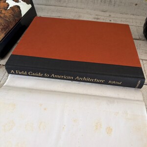 May include: A hardcover book titled A Field Guide to American Architecture by Rifkind. The book features a burnt orange cover with a black spine and gold lettering. The book is resting on a white paper surface with visible light brown stains.