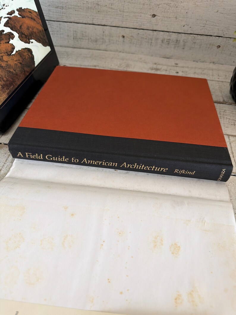May include: A hardcover book titled A Field Guide to American Architecture by Rifkind. The book features a burnt orange cover with a black spine and gold lettering. The book is resting on a white paper surface with visible light brown stains.