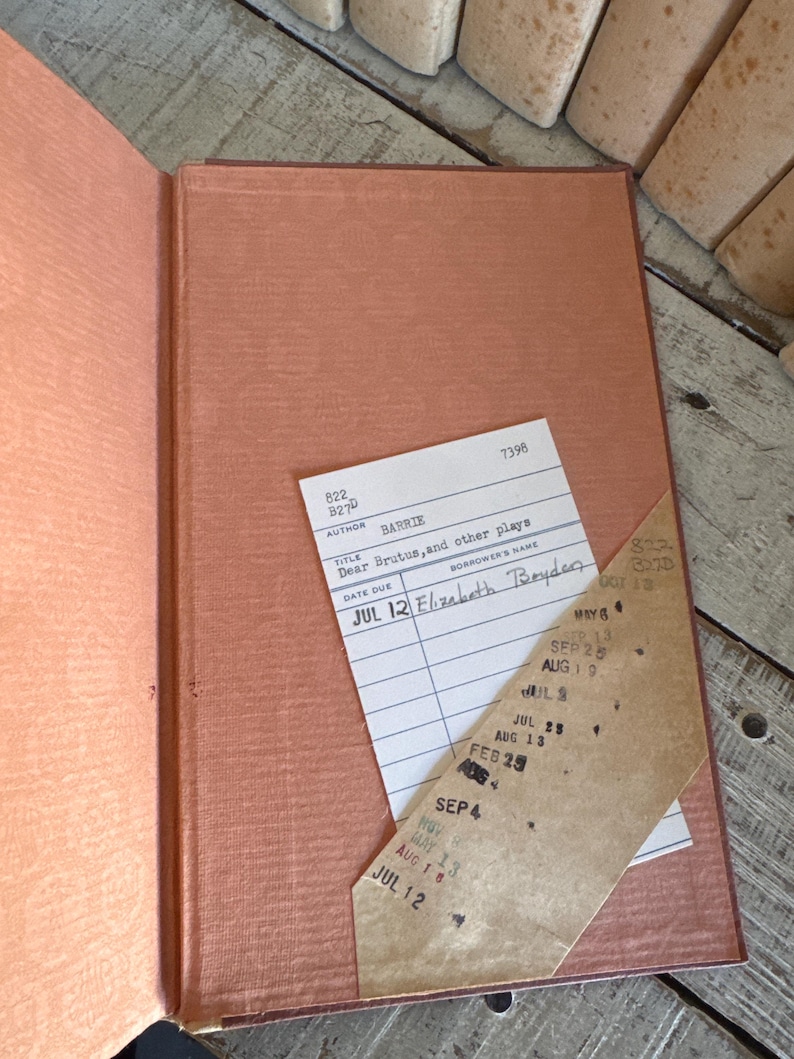 Puede incluir: Libro vintage abierto con una cubierta y p&aacute;ginas de color salm&oacute;n. Una tarjeta de biblioteca est&aacute; metida dentro, con el t&iacute;tulo "Dear Brutus, and other plays" y la fecha "JUL 12". El lomo del libro es visible y las p&aacute;ginas est&aacute;n envejecidas.