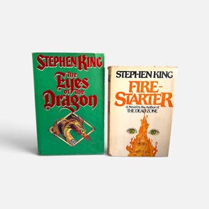 May include: Two hardcover books by Stephen King. The green book is titled The Eyes of the Dragon and features a dragon illustration. The cream-colored book is titled Firestarter and displays a graphic of a face engulfed in orange flames.