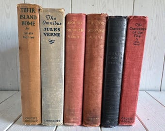 May include: A collection of vintage books by Jules Verne, featuring titles like "Their Island Home" and "Around the World." The books have aged covers in shades of tan, red, and black, with visible text and titles.