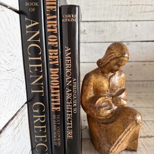 May include: Three black hardcover books titled The Horizon Book of Ancient Greece, The Art of Bev Doolittle, and A Field Guide to American Architecture, positioned next to a small, textured, golden-brown figurine of a seated person holding an object.