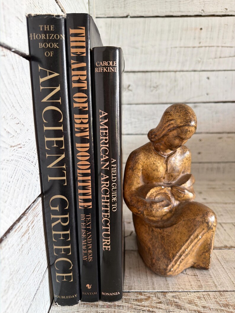 May include: Three black hardcover books titled The Horizon Book of Ancient Greece, The Art of Bev Doolittle, and A Field Guide to American Architecture, positioned next to a small, textured, golden-brown figurine of a seated person holding an object.