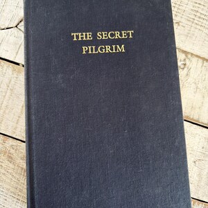 May include: A dark blue hardcover book with the gold title "THE SECRET PILGRIM". The book is closed and lies on a light-coloured, weathered wooden surface.