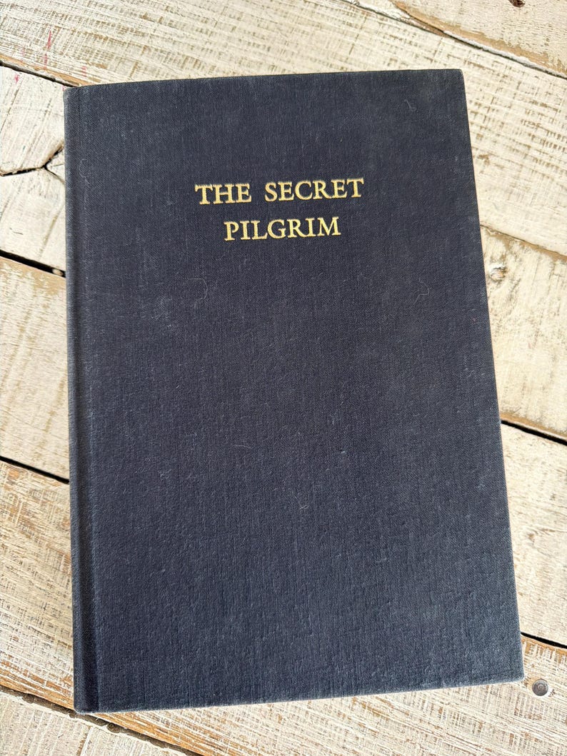 May include: A dark blue hardcover book with the gold title "THE SECRET PILGRIM". The book is closed and lies on a light-coloured, weathered wooden surface.