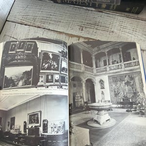 May include: An open book displaying black and white photographs of art galleries and historical buildings. The book rests on a white-painted wooden surface, with dark blue books stacked above.