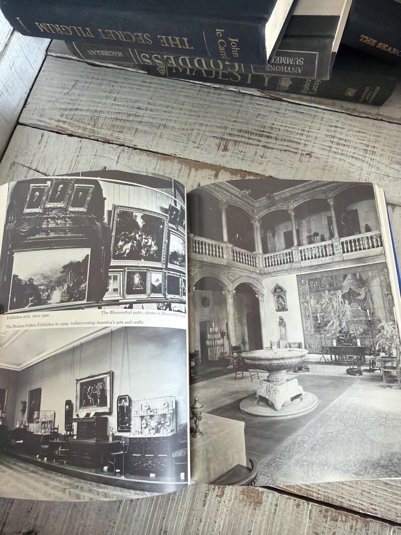 May include: An open book displaying black and white photographs of art galleries and historical buildings. The book rests on a white-painted wooden surface, with dark blue books stacked above.