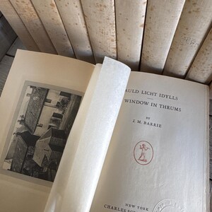 Puede incluir: Un libro abierto con el t&iacute;tulo "Auld Licht Idylls" de J.M. Barrie, publicado por Charles Scribner's Sons en 1919. El libro tiene una ilustraci&oacute;n en blanco y negro y un emblema rojo. Varios libros antiguos est&aacute;n apilados en el fondo.