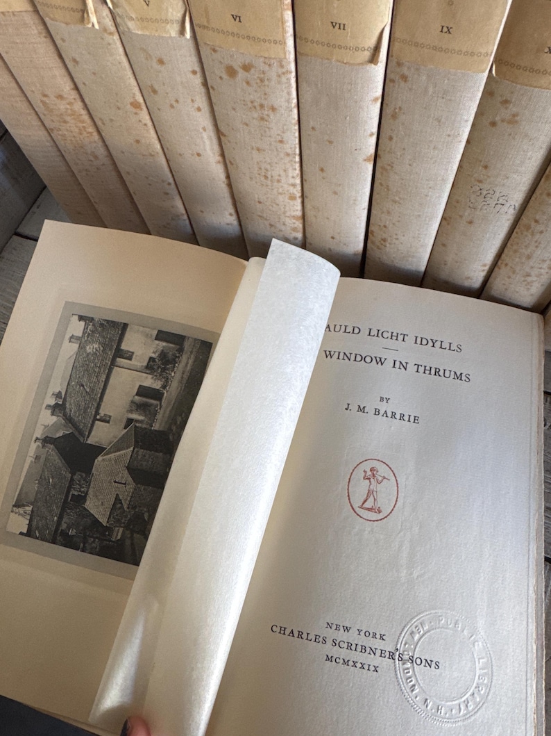 Puede incluir: Un libro abierto con el t&iacute;tulo "Auld Licht Idylls" de J.M. Barrie, publicado por Charles Scribner's Sons en 1919. El libro tiene una ilustraci&oacute;n en blanco y negro y un emblema rojo. Varios libros antiguos est&aacute;n apilados en el fondo.