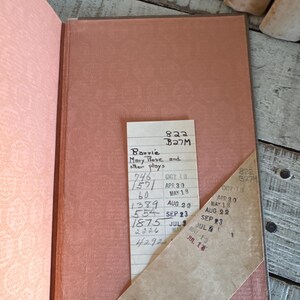 Puede incluir: Libro abierto con una cubierta y p&aacute;ginas de color salm&oacute;n. Una tarjeta de biblioteca con texto manuscrito y fechas selladas est&aacute; insertada. El t&iacute;tulo del libro es "Mary Rose and other plays" de Barrie.