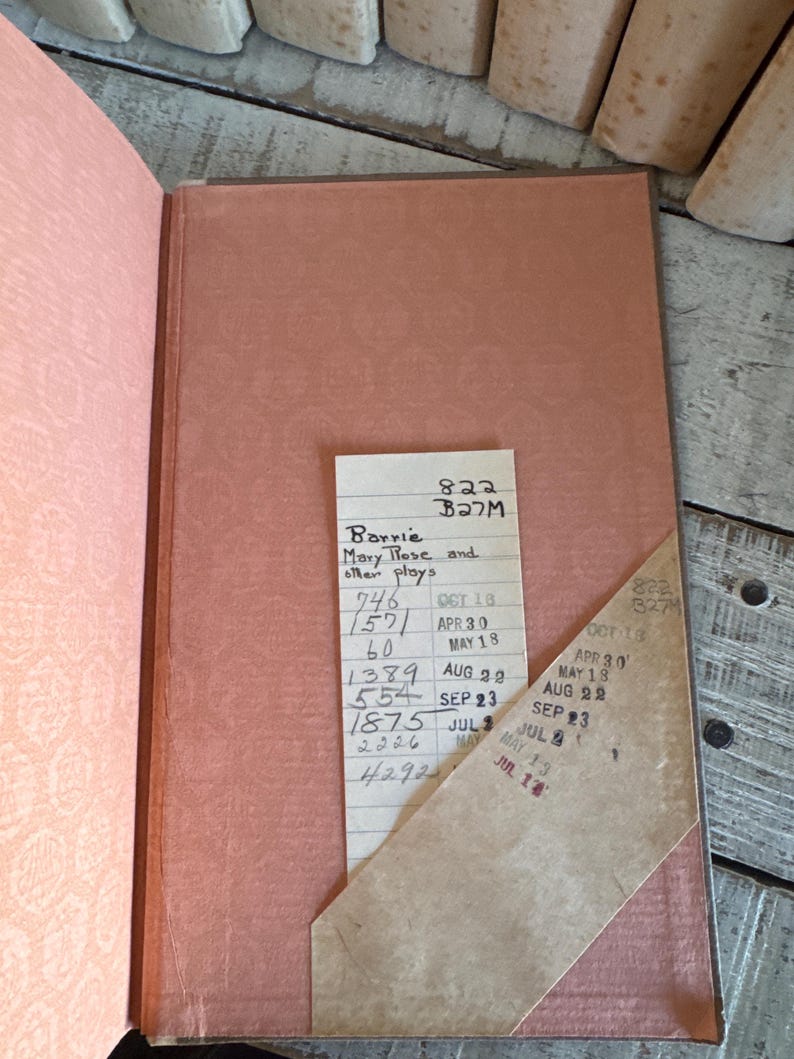 Puede incluir: Libro abierto con una cubierta y p&aacute;ginas de color salm&oacute;n. Una tarjeta de biblioteca con texto manuscrito y fechas selladas est&aacute; insertada. El t&iacute;tulo del libro es "Mary Rose and other plays" de Barrie.