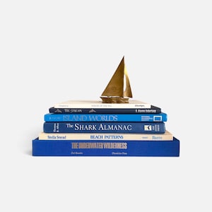 May include: A stack of six books with blue and cream covers, topped with a small brass sailboat figurine. The titles include Voices of the Sea, The Stream, Island Worlds, The Shark Almanac, Beach Patterns, and The Underwater Wilderness.