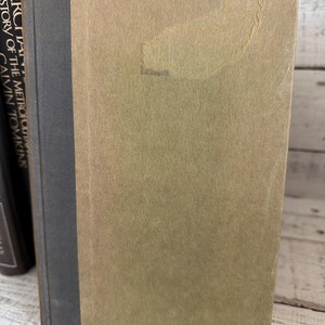 May include: A vintage book with a tan cover and a dark grey spine. The book's cover has a slightly textured appearance with a faded area. Other books are visible in the background, suggesting a collection of literature.