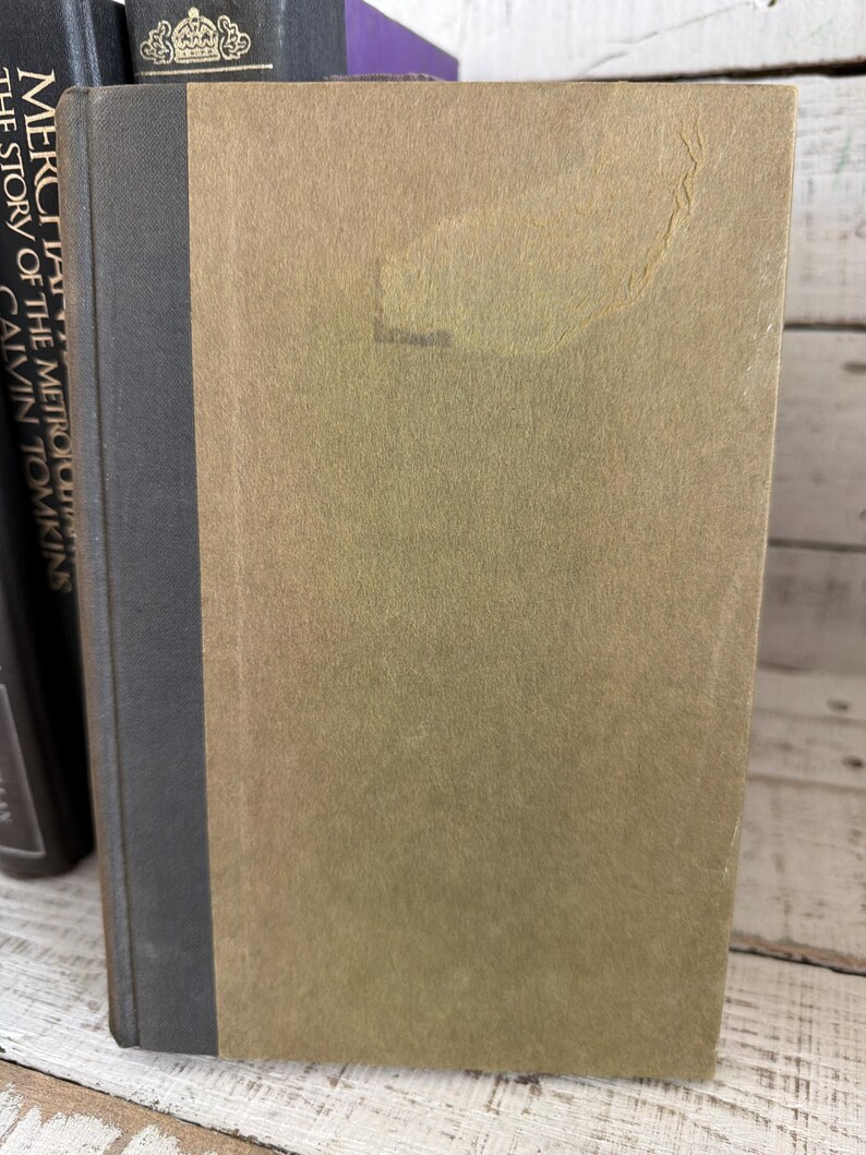 May include: A vintage book with a tan cover and a dark grey spine. The book's cover has a slightly textured appearance with a faded area. Other books are visible in the background, suggesting a collection of literature.