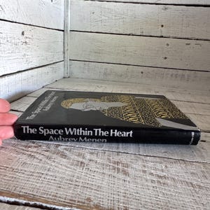 May include: A black hardback book titled "The Space Within The Heart" by Aubrey Menen. The book cover features a black and gold illustration of a person.