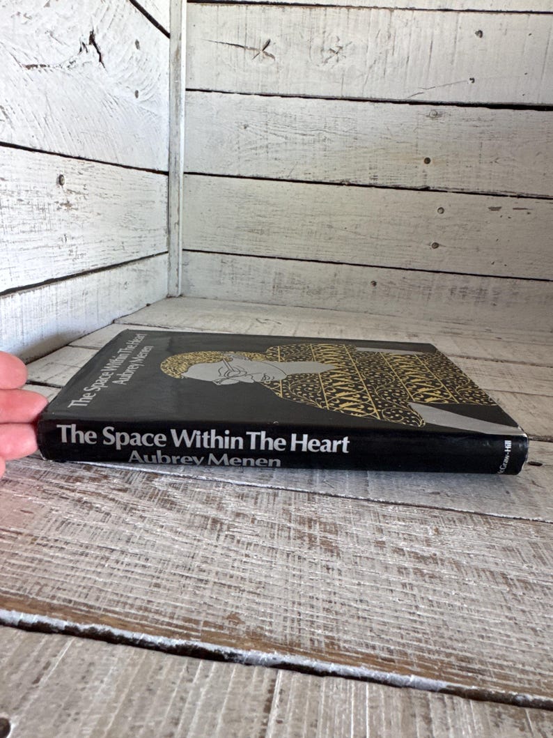 May include: A black hardback book titled "The Space Within The Heart" by Aubrey Menen. The book cover features a black and gold illustration of a person.