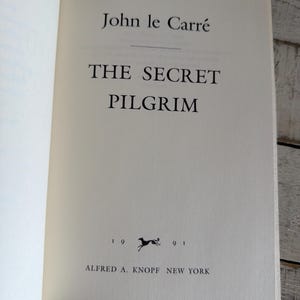 May include: An open book displaying the title "The Secret Pilgrim" by John le Carr&eacute;. The book's pages are off-white, with black text. The publisher's name and year of publication are also visible.