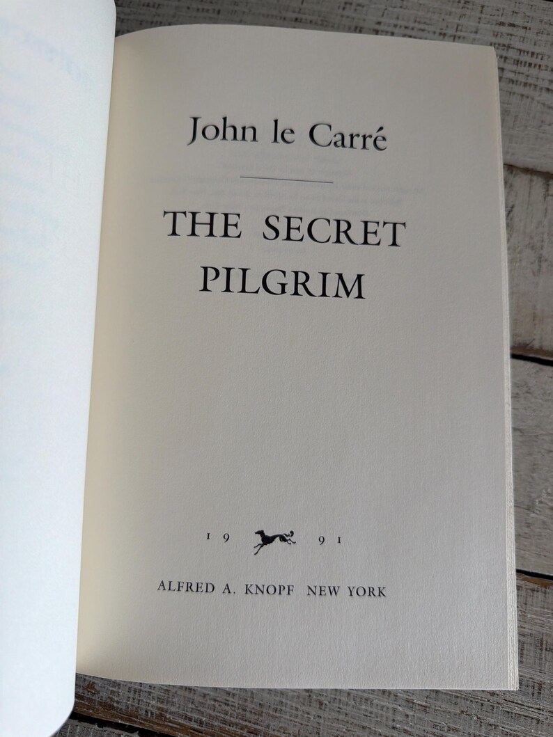 May include: An open book displaying the title "The Secret Pilgrim" by John le Carr&eacute;. The book's pages are off-white, with black text. The publisher's name and year of publication are also visible.