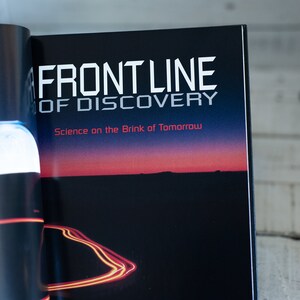 May include: Open book with the title "FRONT LINE OF DISCOVERY" in white, and the subtitle "Science on the Brink of Tomorrow" in red. The background shows a dark landscape with a sunset.