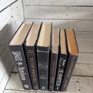 Pu&ograve; includere: Una collezione di libri con copertina rigida e dorsi grigio scuro. I titoli includono "Plum Island" e "To the White Sea". I libri sono disposti su una superficie di legno, con i titoli e gli autori visibili.