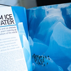 May include: Open book with the title "FROM ICE TO WATER" and text about melting glaciers and ice shelves. The right page shows a blue image of ice formations and penguins. The text "NOT MARCHING ANYMORE? Penguins in Antarctica are imperiled as ongoing melting of polar ice threatens their habitats."