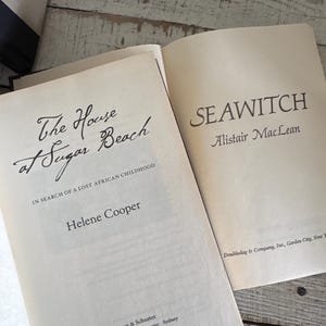 Pu&ograve; includere: Due libri aperti con testo. Il titolo del libro di sinistra &egrave; "The House at Sugar Beach" di Helene Cooper. Il titolo del libro di destra &egrave; "SEAWITCH" di Alistair MacLean. I libri sono su una superficie di legno chiaro e invecchiato.