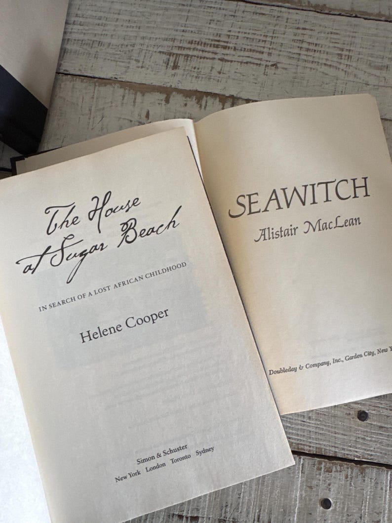 Pu&ograve; includere: Due libri aperti con testo. Il titolo del libro di sinistra &egrave; "The House at Sugar Beach" di Helene Cooper. Il titolo del libro di destra &egrave; "SEAWITCH" di Alistair MacLean. I libri sono su una superficie di legno chiaro e invecchiato.
