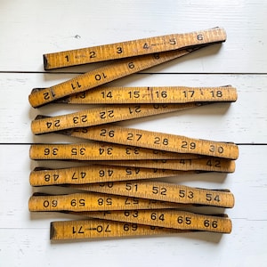 May include: A vintage wooden folding ruler, featuring black numerals and markings. The ruler is a warm, aged yellow color, with visible wear and tear. The ruler extends to 71 inches.