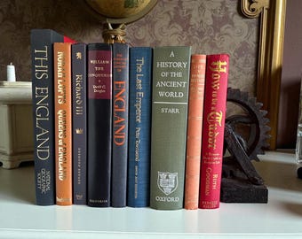 May include: A collection of hardcover books with various titles, including "This England" and "How to be a Tudor." The books are in different colors, such as black, orange, and green, and are arranged on a white surface. A decorative globe and gear are in the background.