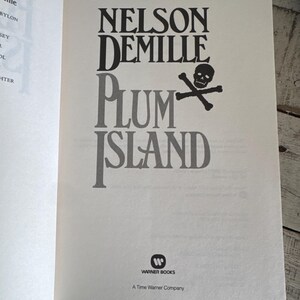 Pu&ograve; includere: Pagina di libro aperta con il titolo "PLUM ISLAND" di Nelson DeMille. Il titolo &egrave; in grandi lettere nere e grigie, con un teschio e ossa incrociate. Il logo Warner Books &egrave; in basso.