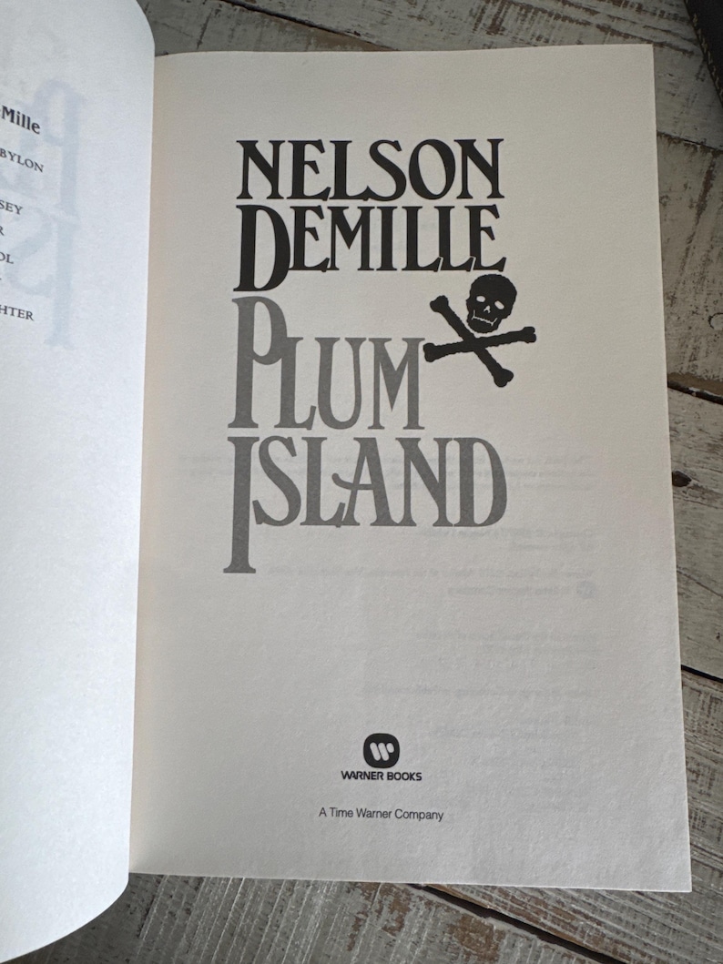 Pu&ograve; includere: Pagina di libro aperta con il titolo "PLUM ISLAND" di Nelson DeMille. Il titolo &egrave; in grandi lettere nere e grigie, con un teschio e ossa incrociate. Il logo Warner Books &egrave; in basso.