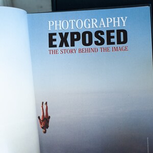 May include: Open book with the title "PHOTOGRAPHY EXPOSED" in white and black text. The subtitle reads "THE STORY BEHIND THE IMAGE" in red. A skydiver in a red suit is falling against a blue sky background.