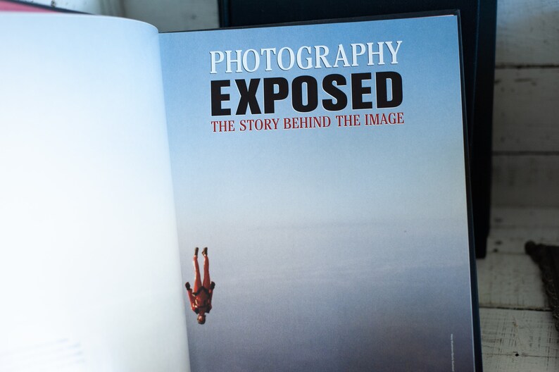 May include: Open book with the title "PHOTOGRAPHY EXPOSED" in white and black text. The subtitle reads "THE STORY BEHIND THE IMAGE" in red. A skydiver in a red suit is falling against a blue sky background.